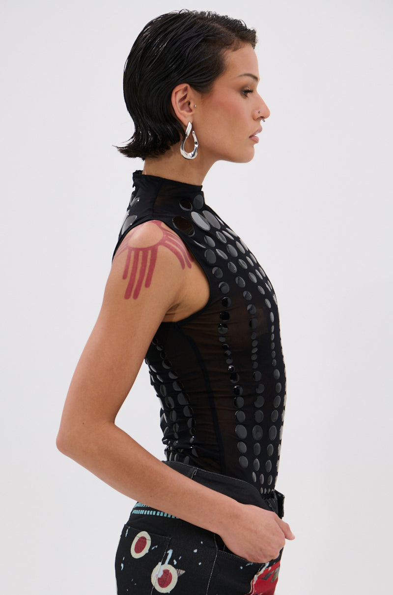 A person with short wet-look hair, hoop earrings, and black pants with a red and white pattern stands in profile against a white background wearing the NOT MY FAULT SLEEVELESS BODYSUIT IN BLACK.