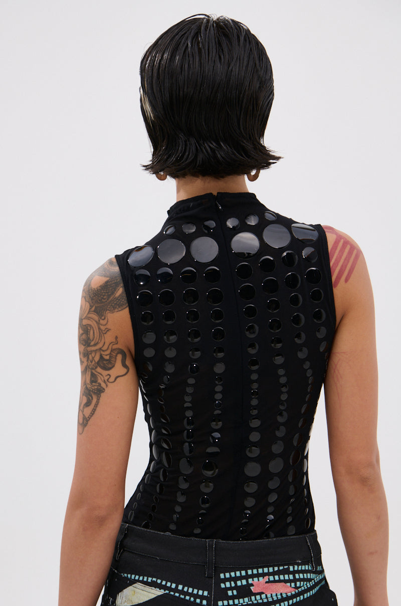 A person with short, dark, wet-look hair models the NOT MY FAULT SLEEVELESS BODYSUIT IN BLACK from the back, featuring cheeky circular cutouts, shiny accents, and revealing a detailed tattoo on their left arm.