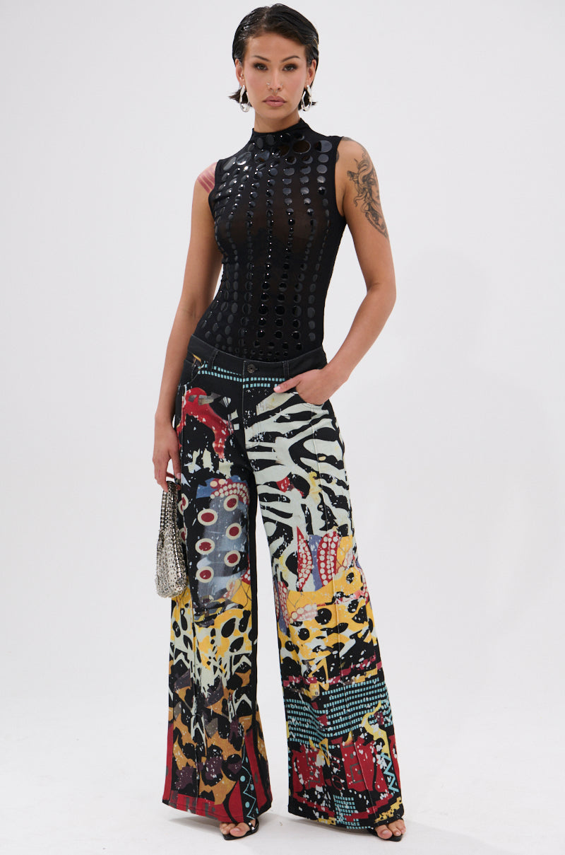 A person with short hair and tattoos stands against a white background, wearing the NOT MY FAULT SLEEVELESS BODYSUIT IN BLACK—sheer black mesh—paired with colorful wide-leg pants and holding a small handbag.