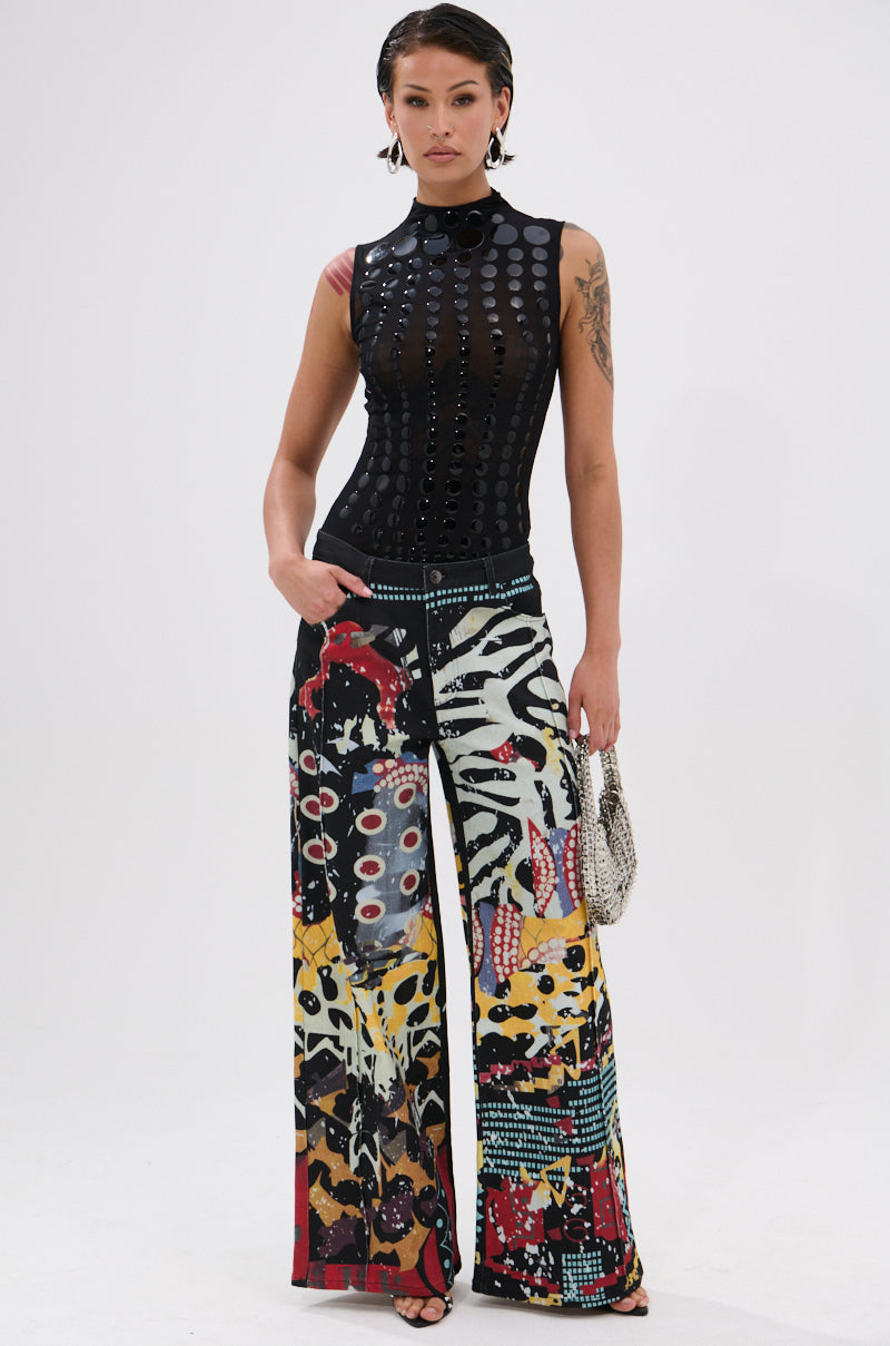 A woman poses against a white background wearing the NOT MY FAULT SLEEVELESS BODYSUIT IN BLACK, a semi-sheer mesh top with shiny dots, paired with high-waisted, wide-leg patchwork pants and a small silver purse.