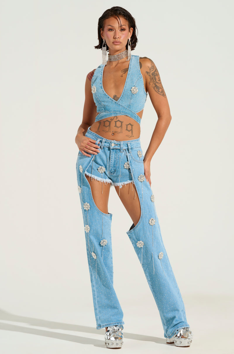 A person poses confidently in the DIVALICIOUS DENIM CHAPS, showcasing its halter top and distinct open-front jeans adorned with rhinestone embellishments. They complete the look with silver platform shoes, revealing tattoos on their arms and torso against a plain backdrop.