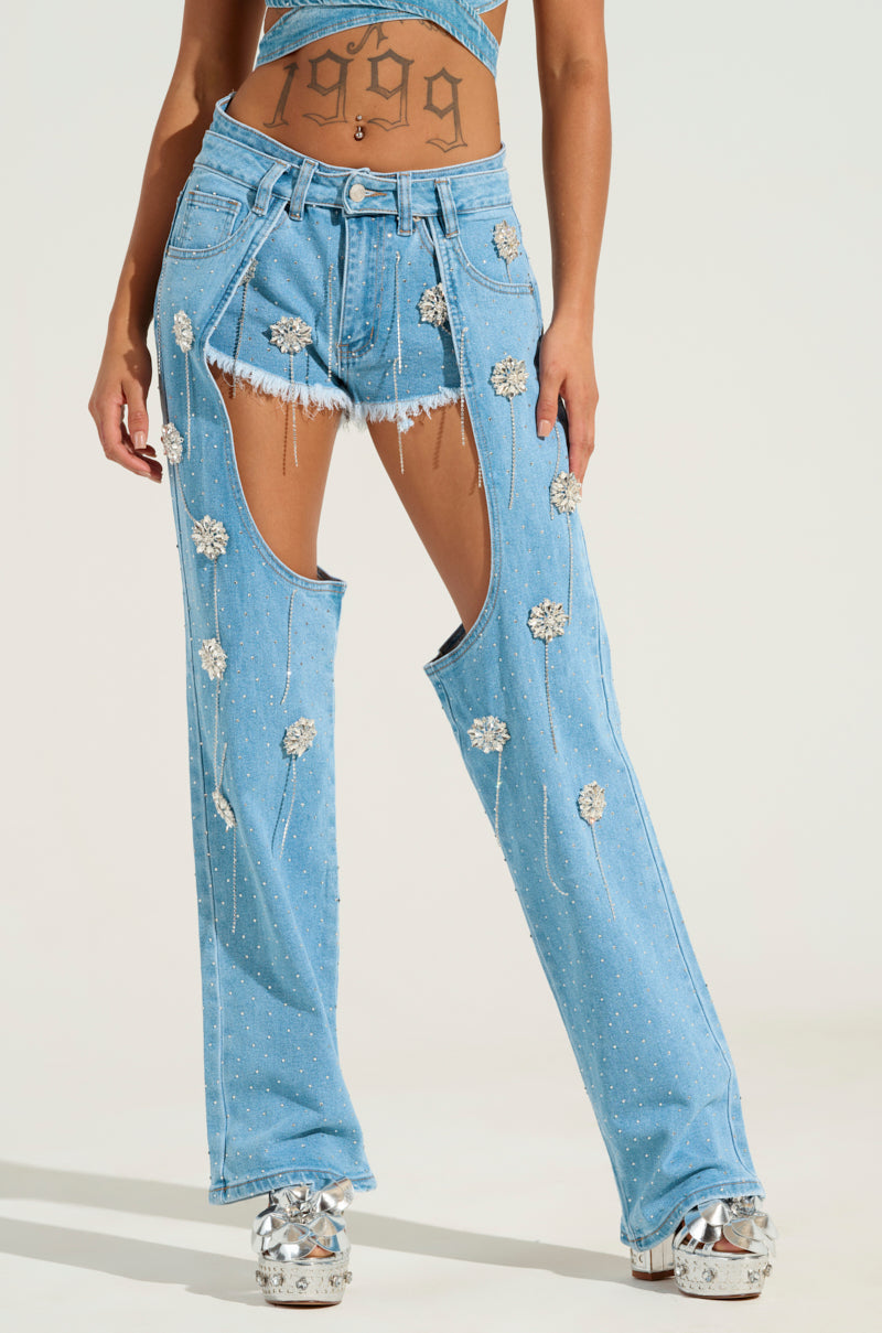 A person wearing the distinctive DIVALICIOUS DENIM CHAPS, featuring high-rise fit with cutouts and floral embellishments. The ensemble is completed with platform shoes, showcasing a tattooed stomach. The design also includes rhinestone details and a belt on the upper section.