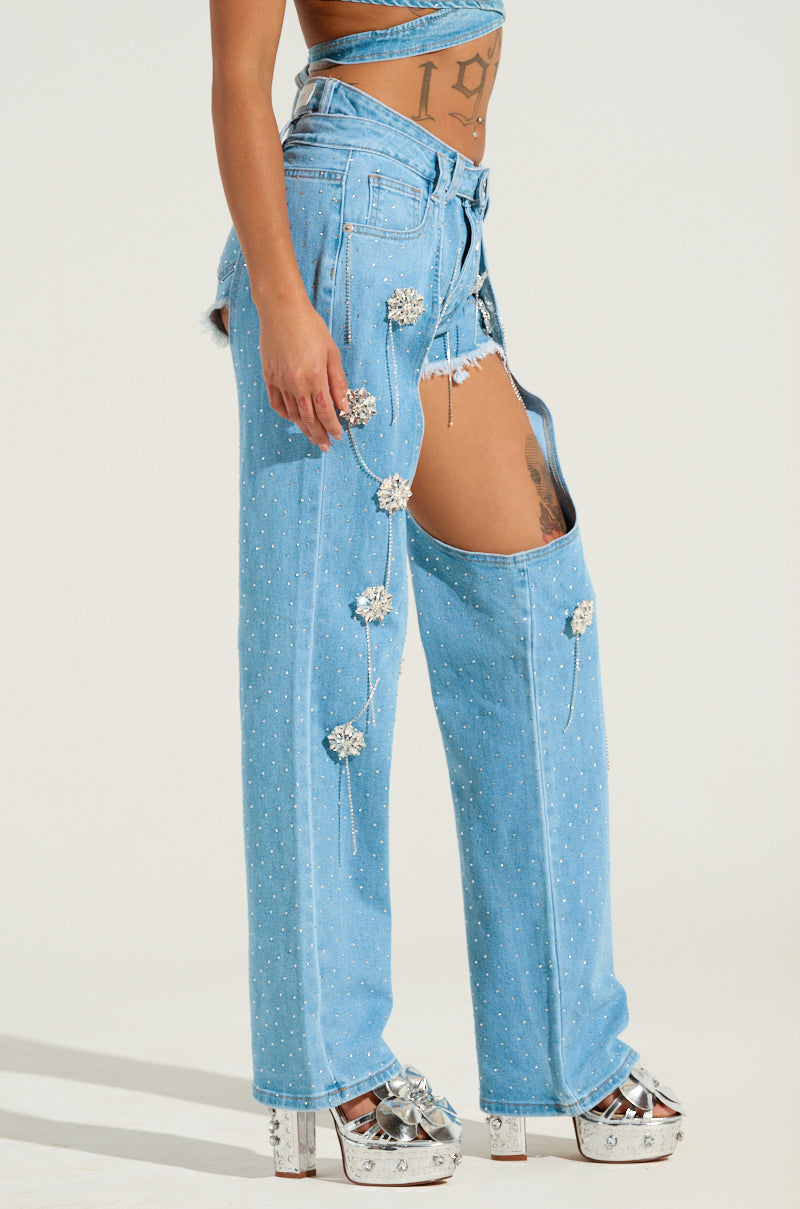 A person is wearing the DIVALICIOUS DENIM CHAPS, featuring a light blue wash with floral embellishments and large thigh cutouts. The chaps are combined with high-platform silver heels, and as they stand sideways, a tattoo on their torso is visible.