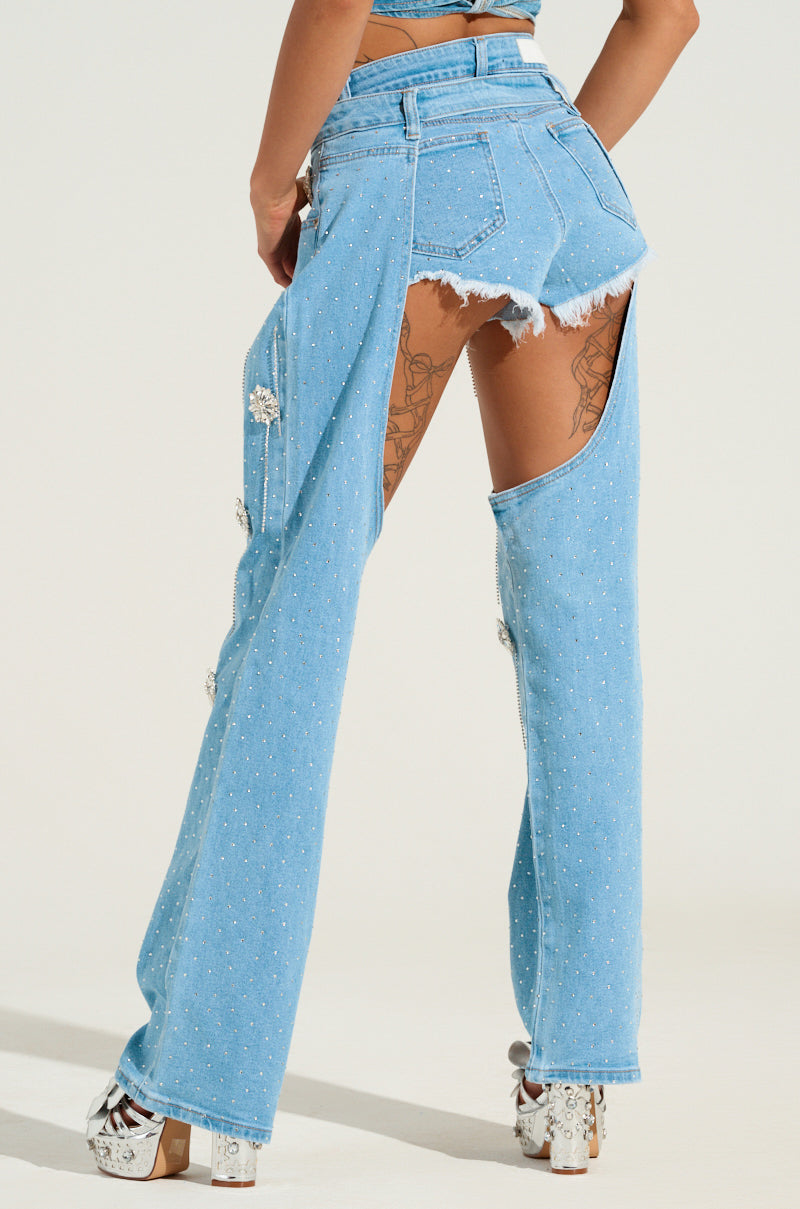 A person wearing the distinctive DIVALICIOUS DENIM CHAPS stands with their back to the camera. The chaps feature intricate small studs and striking open slits along the sides, revealing the thighs, and are ideally paired with platform heels. Tattoos can be seen through on the legs.