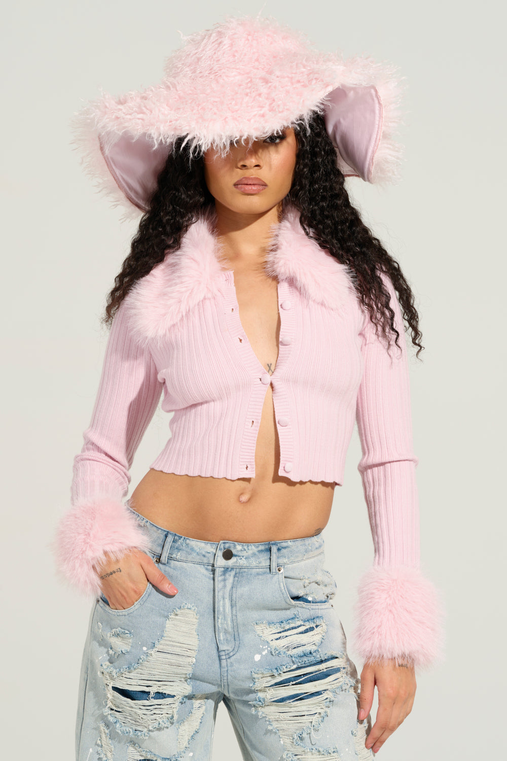 A person wears a fluffy pink hat and an Angel Eyes Fur Trim Cardigan in Pink, featuring a feathered trim at the collar and cuffs. They also sport distressed blue jeans, with their long, curly hair flowing underneath the hat.