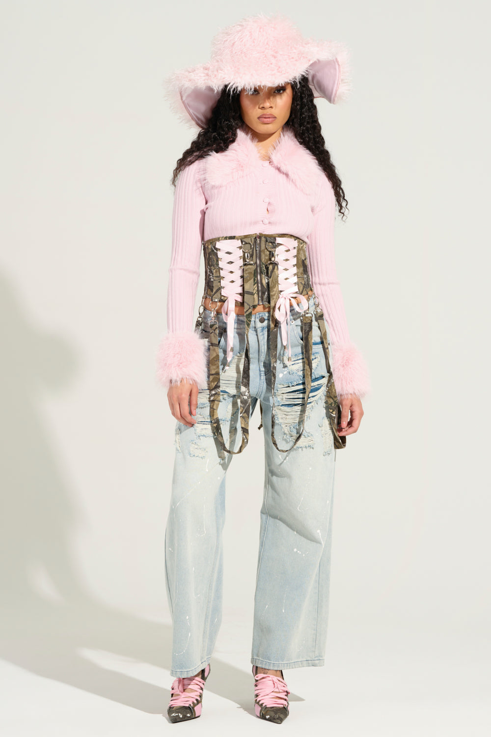 A person wearing a fluffy pink wide-brimmed hat, an Angel Eyes Fur Trim Cardigan in Pink over a camouflage corset with laces, light blue distressed jeans, and pink lace-up shoes stands against a plain background.