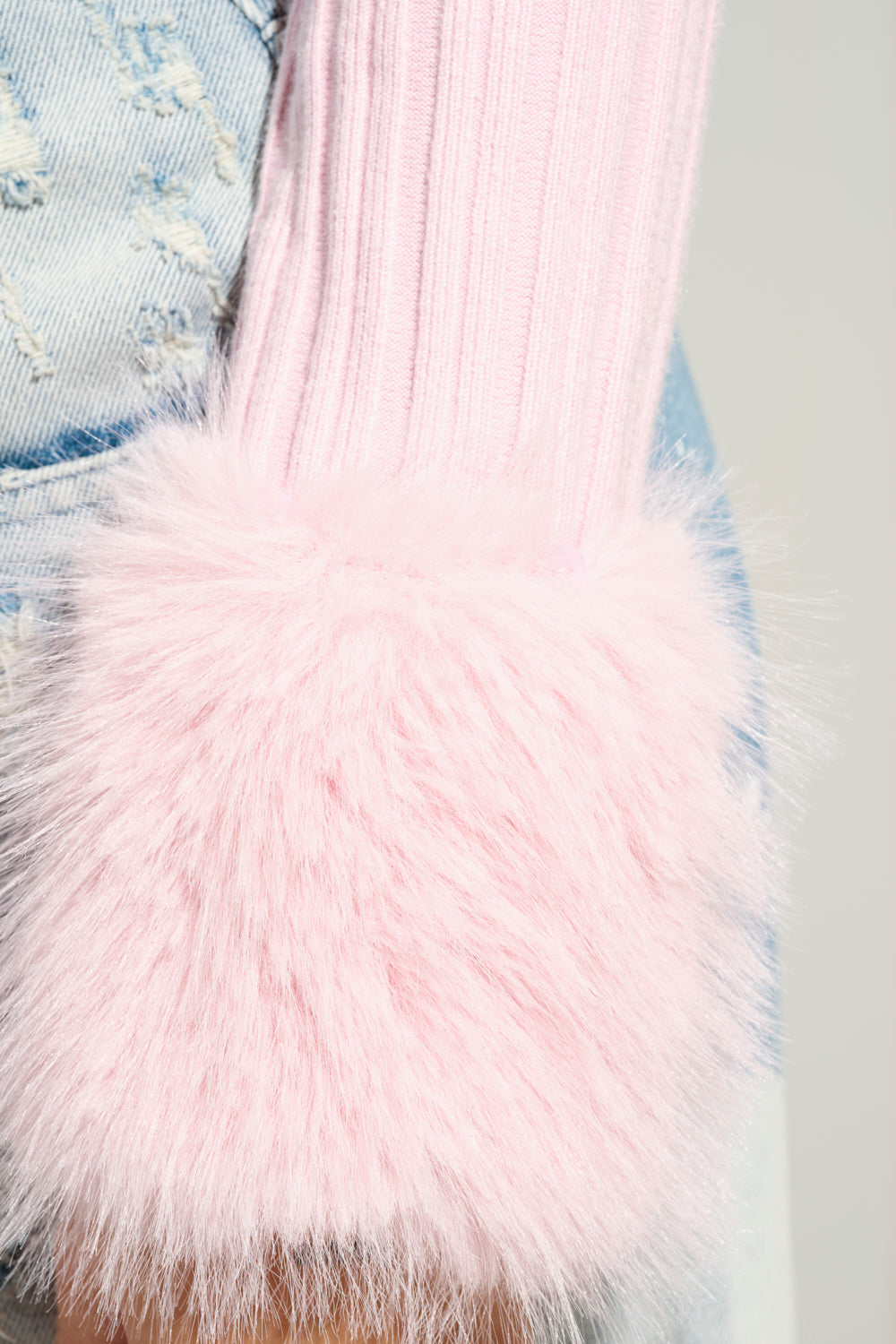 A close-up of a person wearing an ANGEL EYES FUR TRIM CARDIGAN IN PINK, featuring ribbed knit fabrication and fluffy faux fur cuffs. The cardigan is paired with light blue, distressed denim jeans against a soft, neutral background.