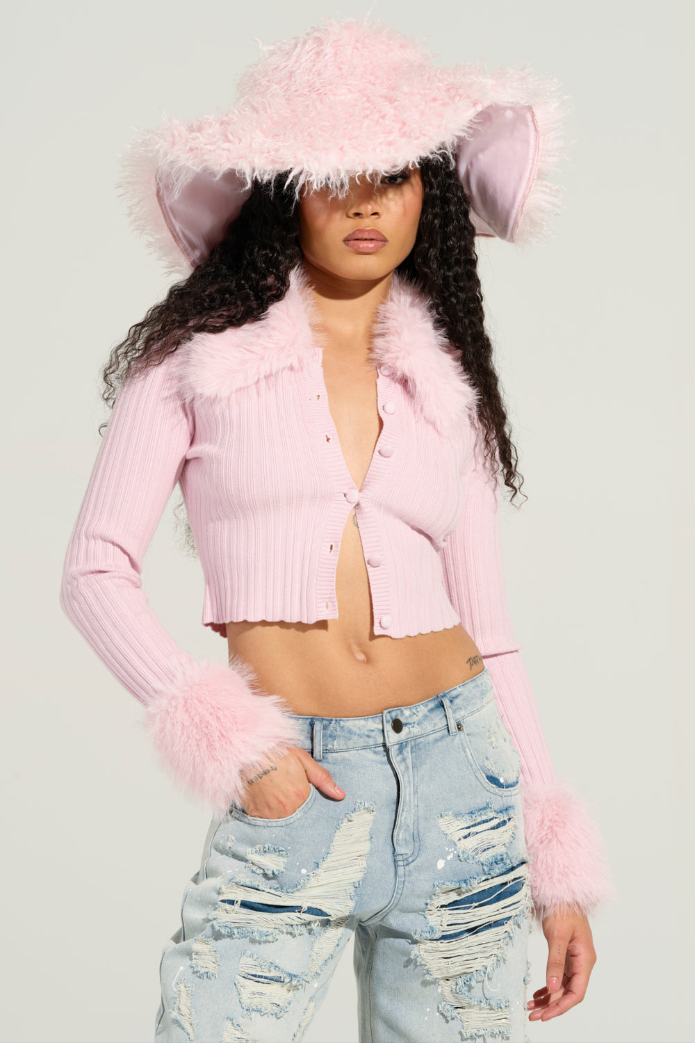 A person poses confidently wearing the ANGEL EYES FUR TRIM CARDIGAN IN PINK and a large, fluffy pink hat. They have long, curly hair and complement their look with light blue, distressed jeans against a plain, light-colored background.