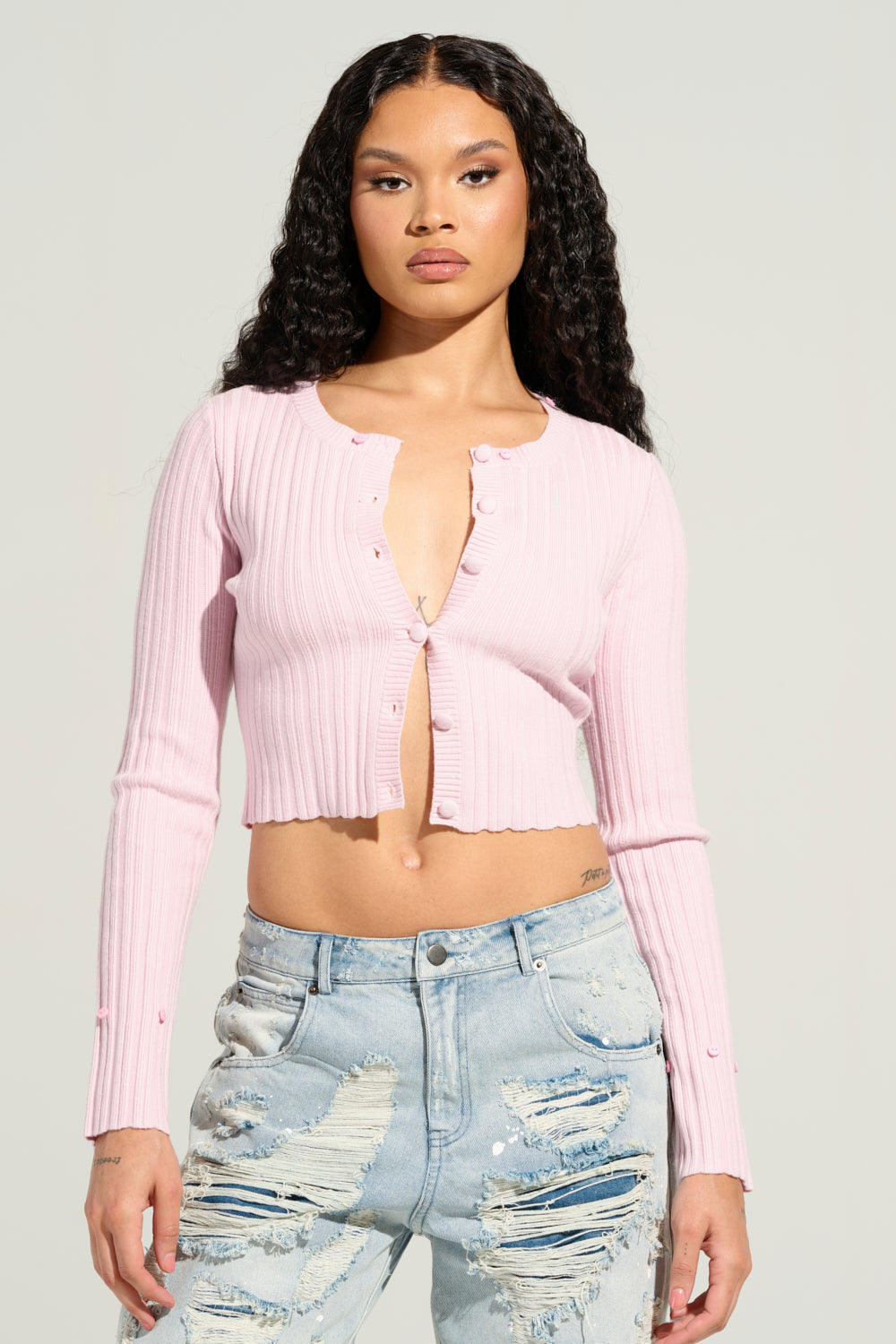 A person with long, curly hair stands confidently facing forward, wearing the Angel Eyes Fur Trim Cardigan in Pink paired with light blue distressed jeans against a plain background.