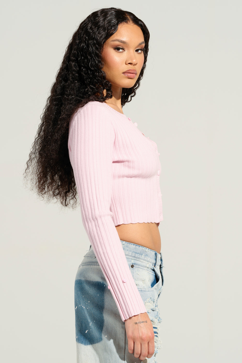 A woman with long, curly hair stands sideways, exuding confidence in an ANGEL EYES Fur Trim Cardigan in Pink and light blue distressed jeans against a plain, neutral background.