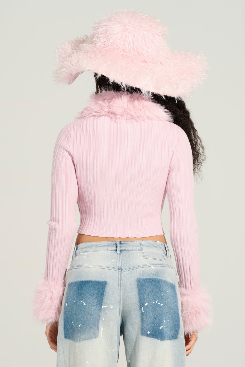 A person with long curly hair is wearing a cozy pink hat and the Angel Eyes Fur Trim Cardigan in Pink, which features a charming faux fur collar. They complement this look with light blue jeans decorated with white paint splatters, facing away from the camera.