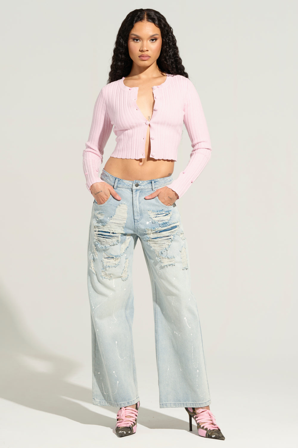 A woman with long curly hair stands confidently, wearing the Angel Eyes Fur Trim Cardigan in pink and ripped light blue jeans. Her hands are in her pockets, and a pair of pink strappy heels completes her outfit. The background is plain and light-colored.
