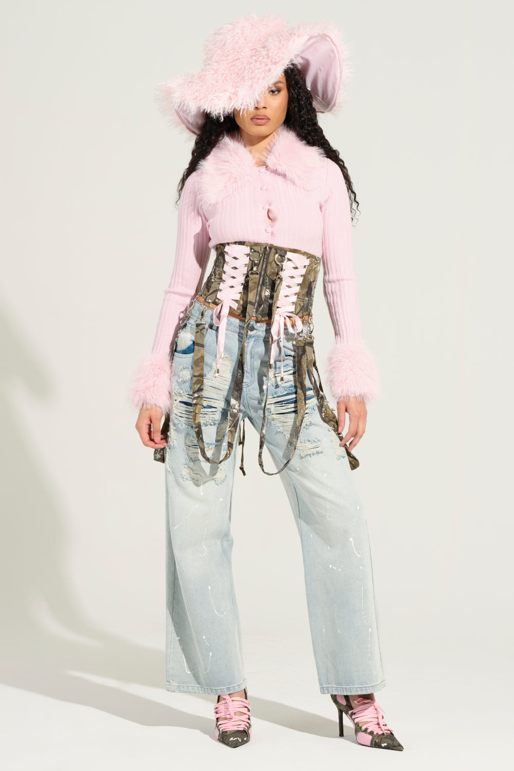 A person stands confidently in a large pink fuzzy hat, the Angel Eyes Fur Trim Cardigan in Pink featuring a faux fur collar, a corset-style top, distressed wide-leg jeans, and pink lace-up heels. This combination of ribbed knit fabrication and bold textures seamlessly merges vintage and modern styles with flair.