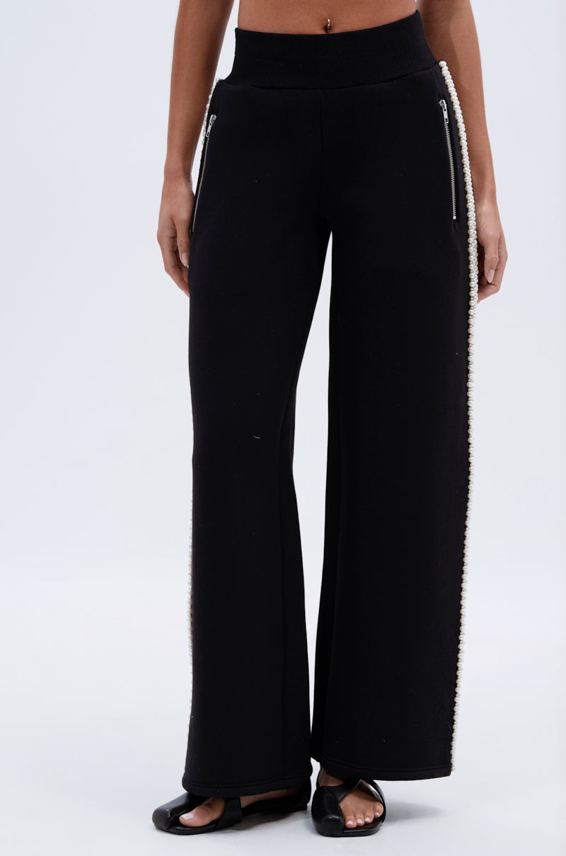 A person wearing the PEARL GIRL JOGGER—black wide-leg sweatpants with zipper pockets and pearl details along the side seams—paired with black open-toe sandals. The photo is cropped from the shoulders down.