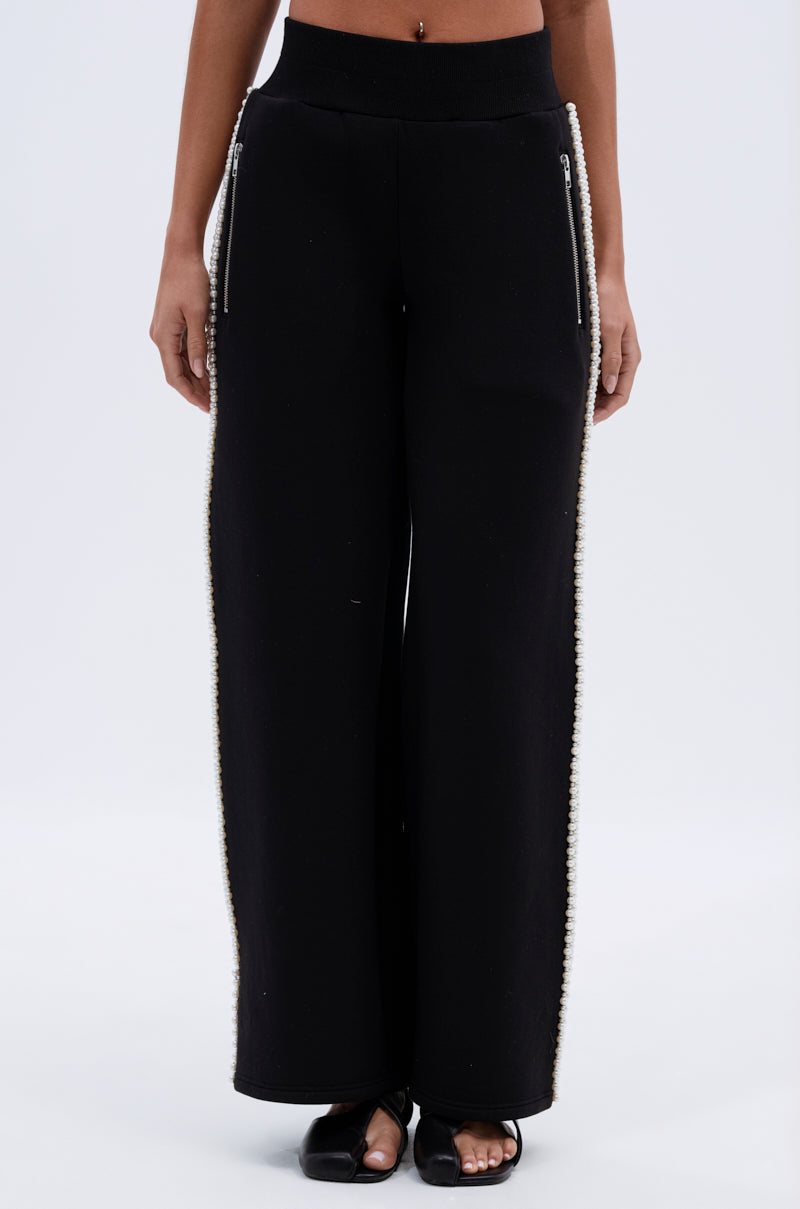 A person wears the PEARL GIRL JOGGER, featuring high rise, wide leg fit, pearl embellishments along side seams, zippered pockets, and black sandals. The stylish joggers stand out against a plain white background.