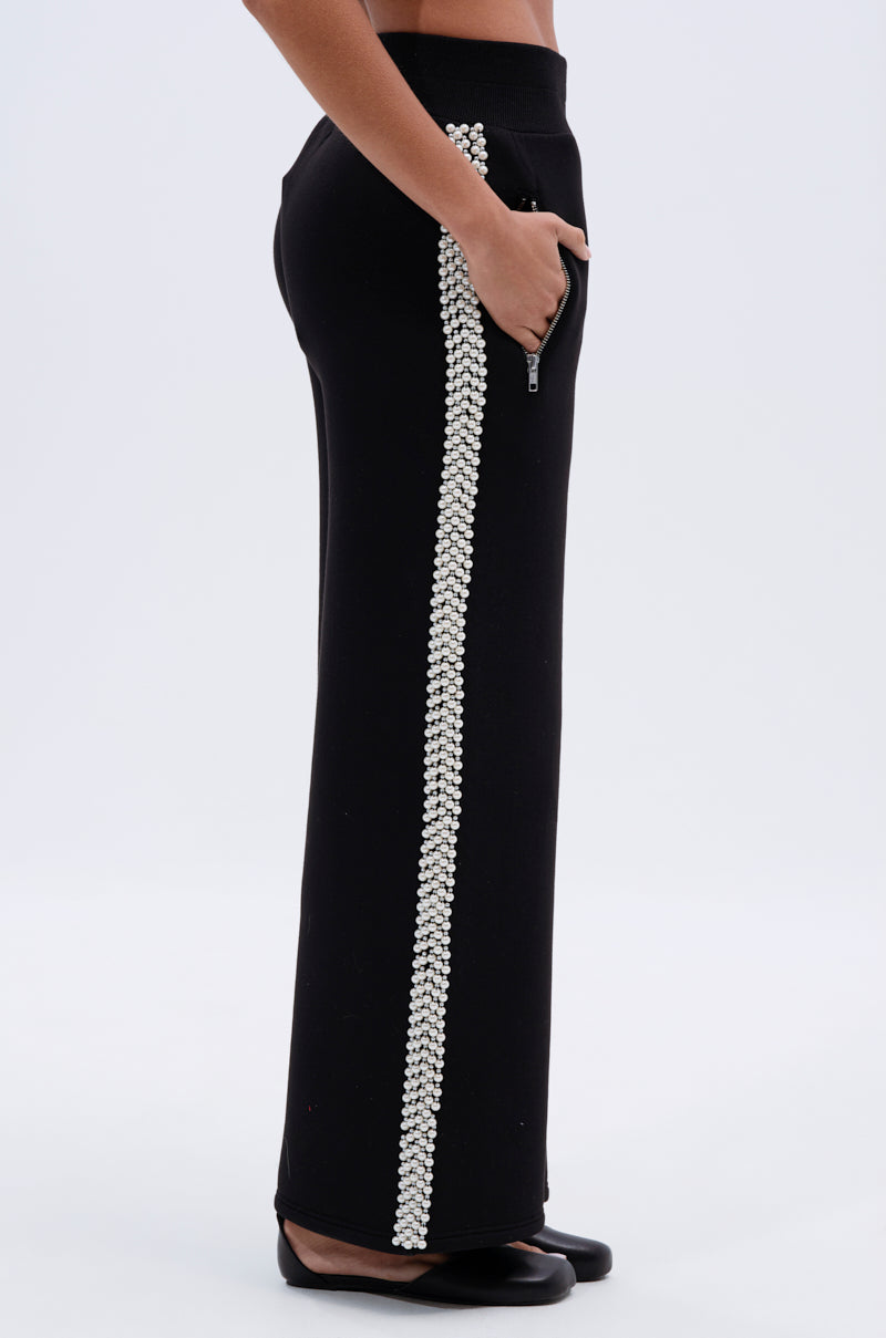 A person wearing PEARL GIRL JOGGER pants—black, high rise, wide leg with a vertical line of silver embellishments on the side seam—stands sideways with one hand in their pocket.