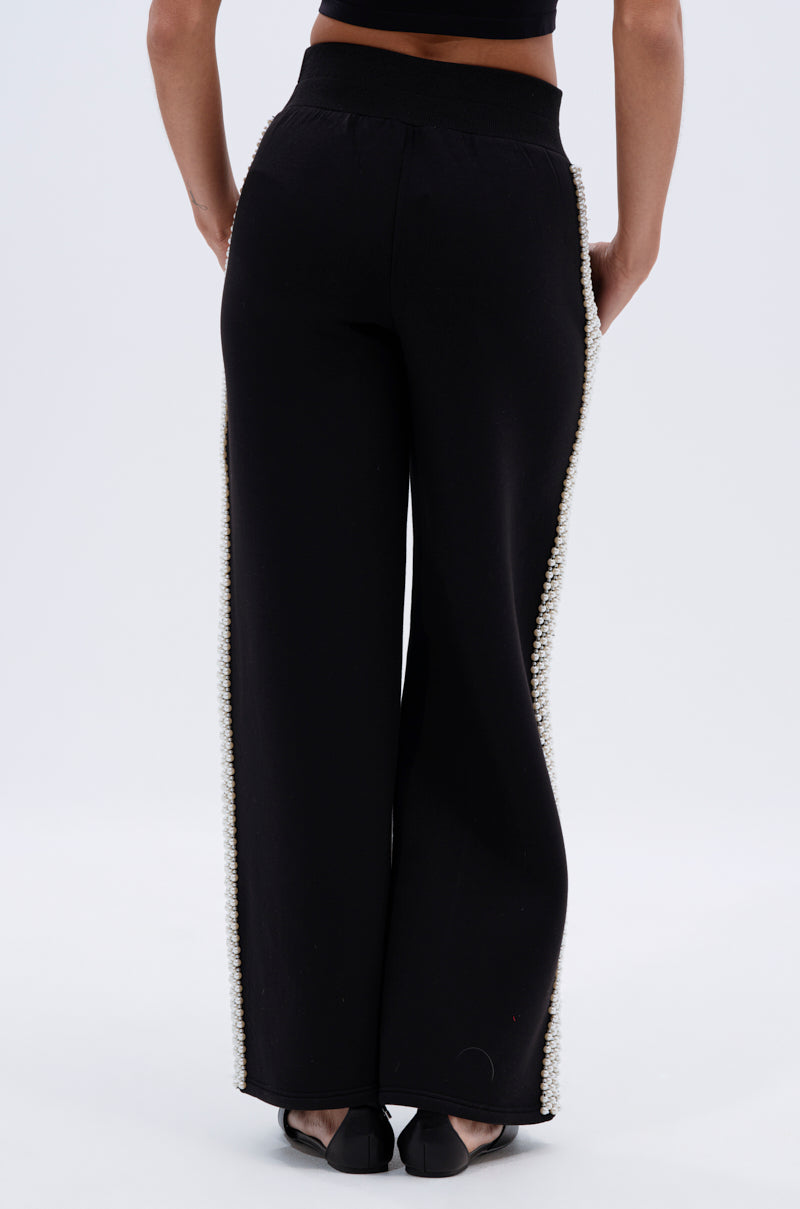 A person wearing the PEARL GIRL JOGGER, black high-rise jogger pants with pearl embellishments along the side seams, is seen from the back against a plain light background.