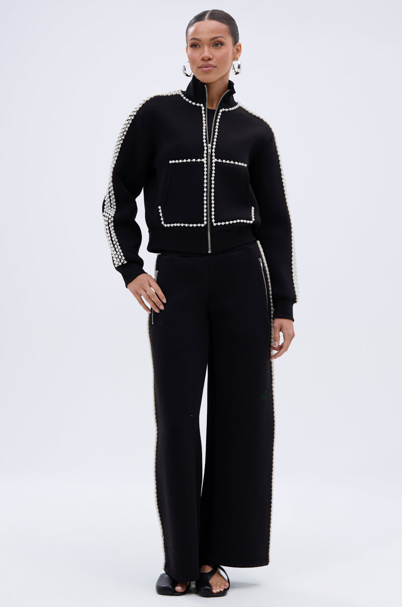 A woman wears the PEARL GIRL JOGGER, wide leg sweatpants with white bead detailing along the edges and seams, paired with a black zip-up jacket, hoop earrings, pulled-back hair, and open-toe black shoes.