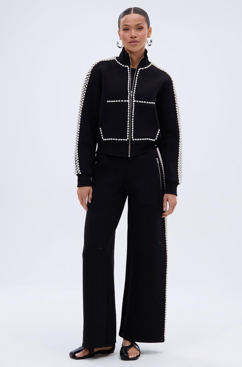 A woman stands against a plain background wearing the PEARL GIRL JOGGER set—a black zip-up jacket and jogger pants featuring white pearl embellishments along the edges—paired with black open-toe sandals.