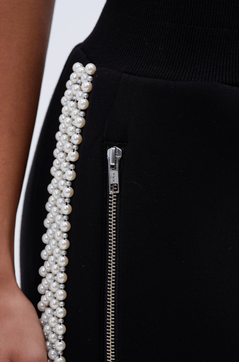 Close-up of the PEARL GIRL JOGGER in black, showcasing a vertical silver zipper and a decorative row of white pearls along the side. A hand rests near the fabric's edge.