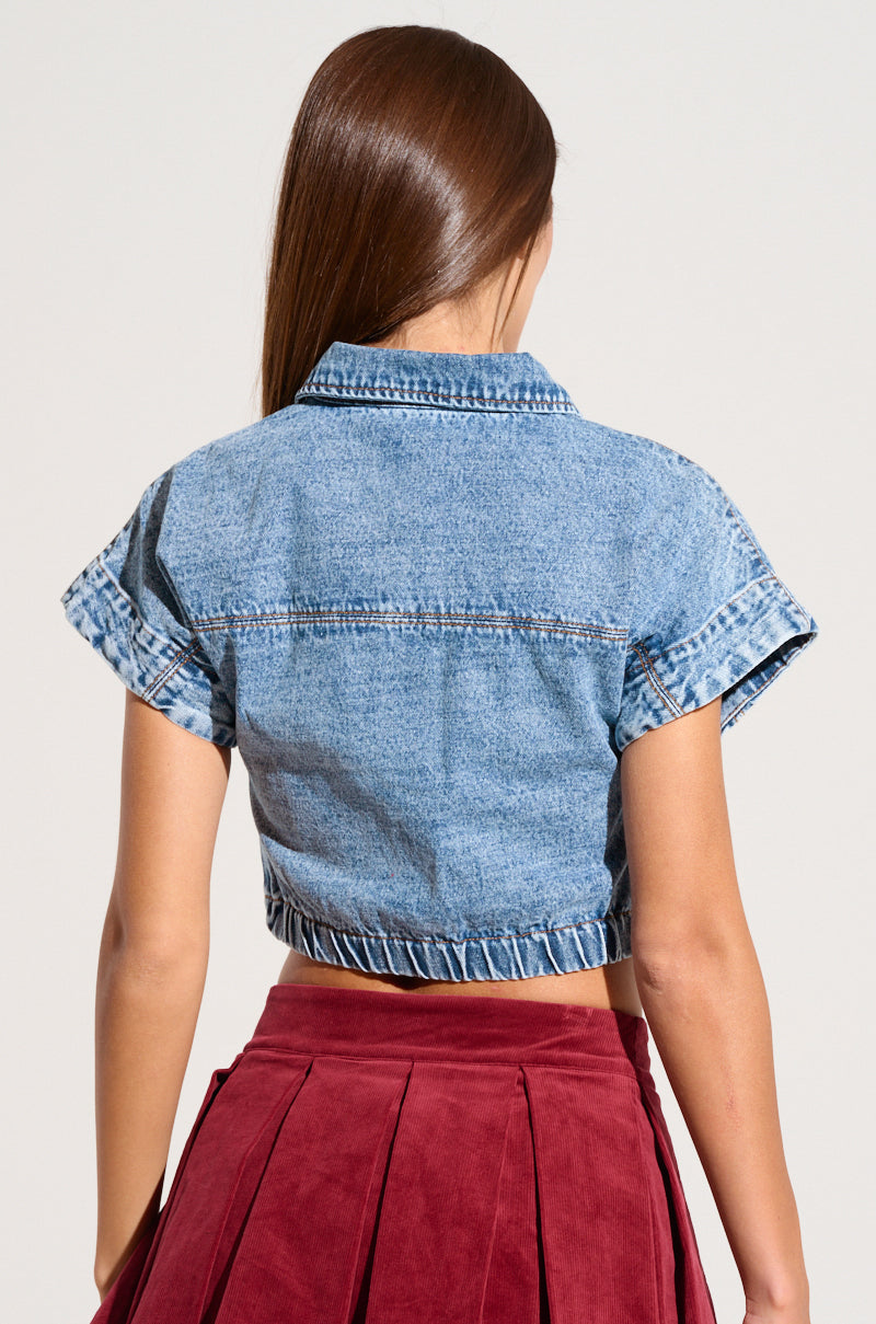 A person with long brown hair is shown from the back. They are wearing the FOREVER THAT GIRL DENIM BUTTON UP SHIRT, a short-sleeved, blue wash denim jacket with a collar, styled like a crop top, and a high-waisted, pleated burgundy skirt. The background is plain and neutral.