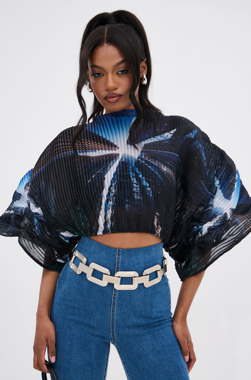 A woman with long dark hair in a ponytail wears the GALAXY BLOUSE, an oversized pleated top with batwing sleeves and a blue and white abstract pattern, paired with high-waisted blue jeans and a chunky silver chain belt.