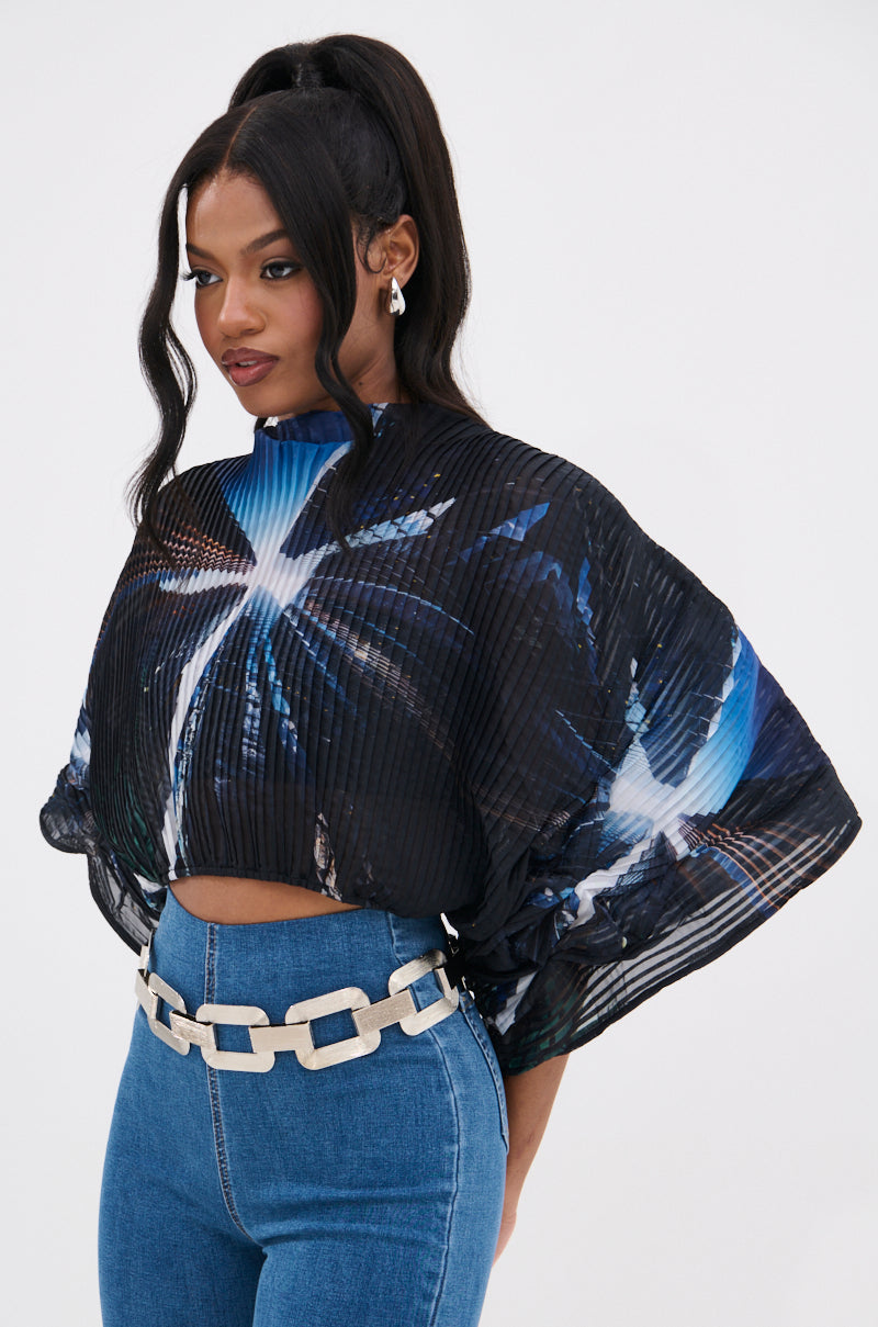 A woman wears the GALAXY BLOUSE—a black pleated top with blue and white abstract patterns—paired with blue high-waisted jeans, a chunky white chain belt, pearl earrings, and her hair in a high ponytail.