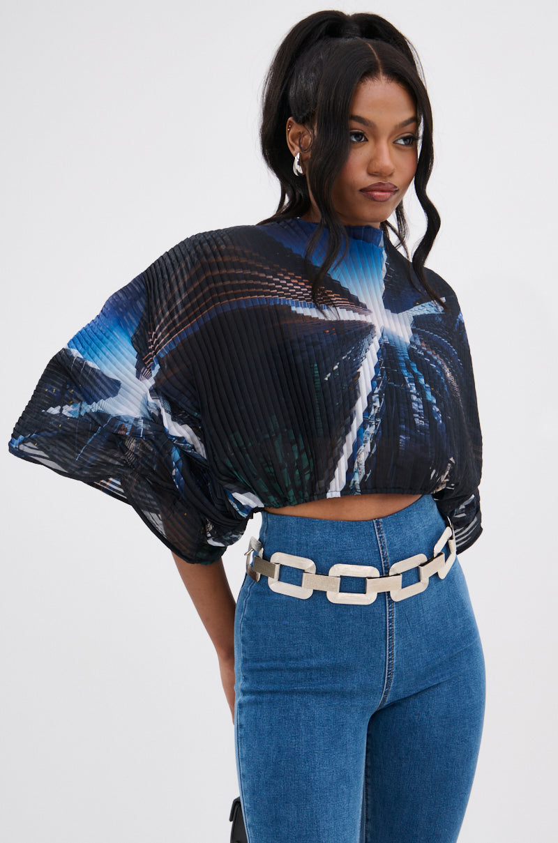 A woman stands against a plain white background wearing the GALAXY BLOUSE—an avant-garde, pleated high-neck top with a blue-and-black galaxy print—paired with high-waisted blue jeans, a chunky silver chain belt, and silver earrings.