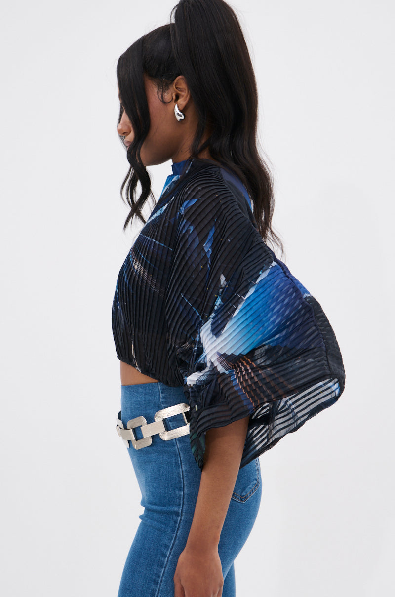A woman with long, wavy hair in a high ponytail models the GALAXY BLOUSE with batwing sleeves, styled with blue jeans, a white chain-link belt, and silver earrings, posing in profile against a plain backdrop.