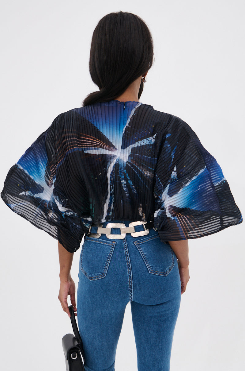 A person with long dark hair wears the GALAXY BLOUSE, featuring blue and white abstract patterns, paired with high-waisted blue jeans and a chunky silver belt. The photo is taken from behind against a plain white background.