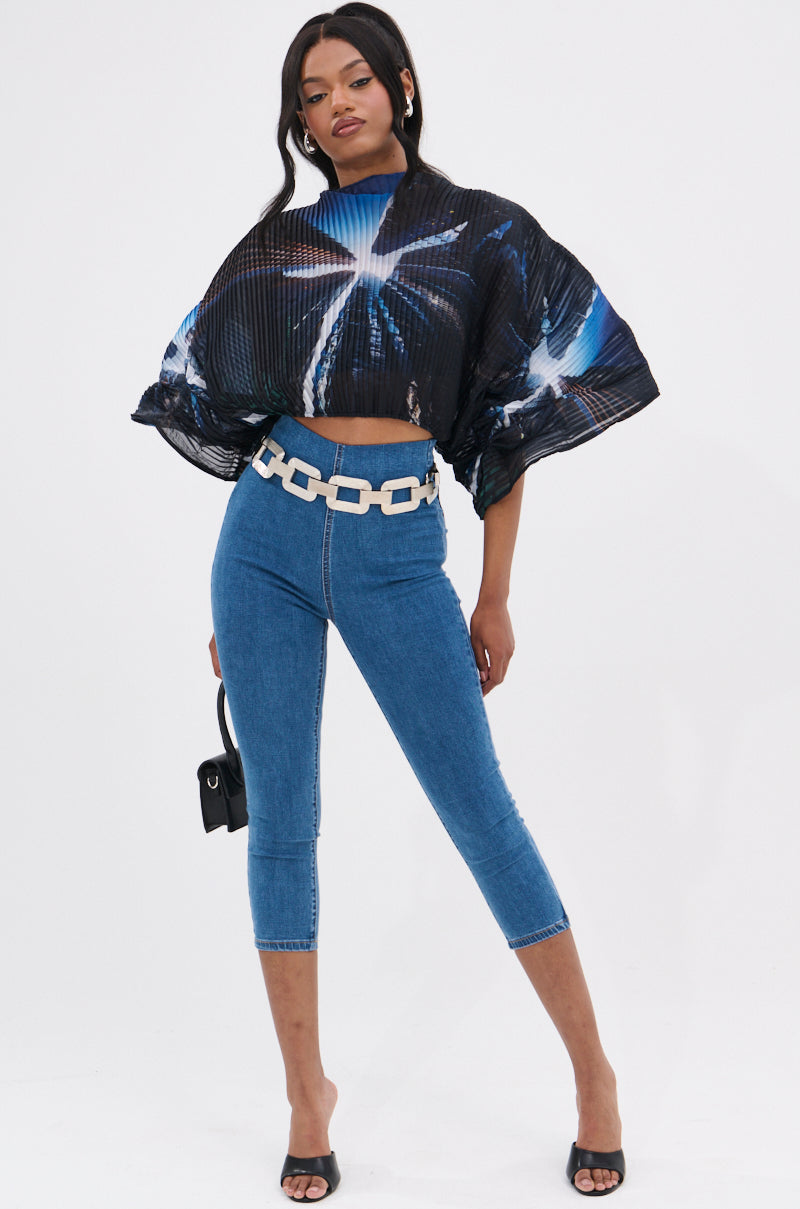A woman poses in the GALAXY BLOUSE, a black crop top with a blue and white city print, paired with high-waisted blue skinny jeans, a chunky white belt, black heeled sandals, and carrying a small black purse.
