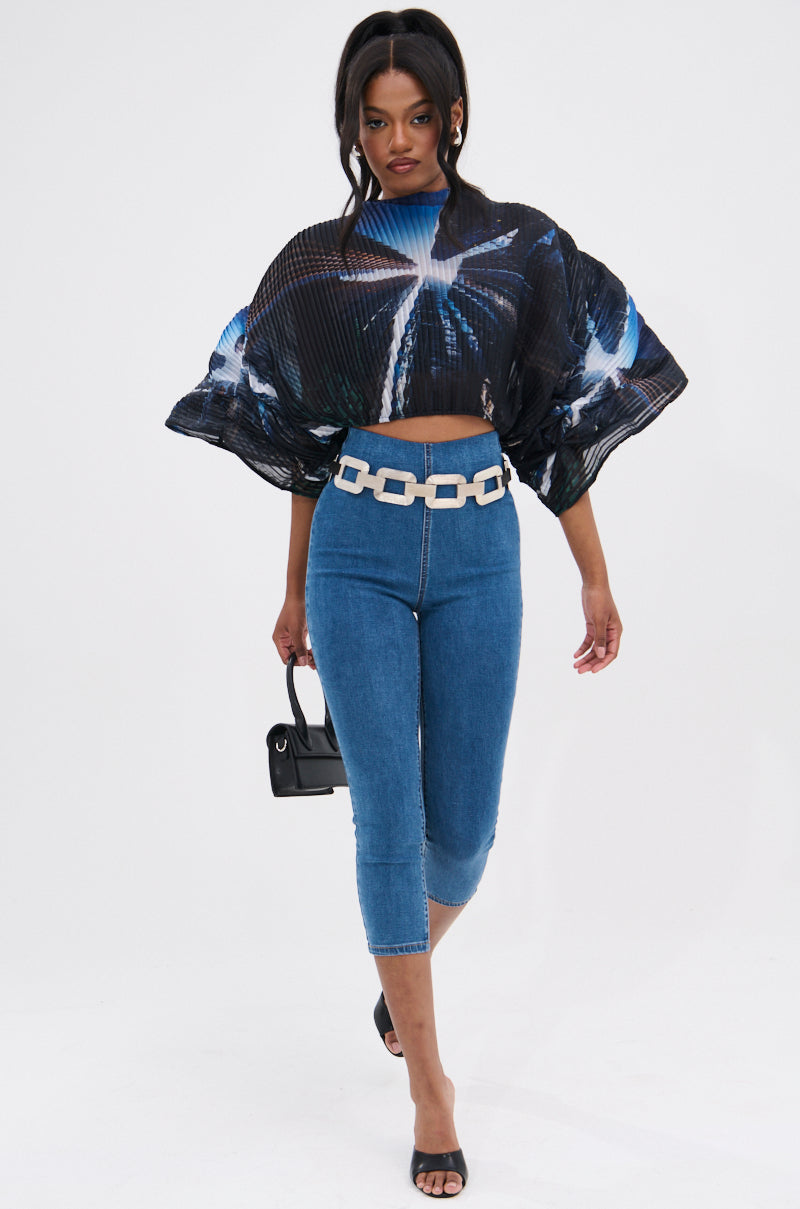 Woman wearing the GALAXY BLOUSE with a blue and white abstract pattern, fitted blue capri jeans, a chunky white belt, black heeled sandals, and carrying a small black handbag walks toward the camera.