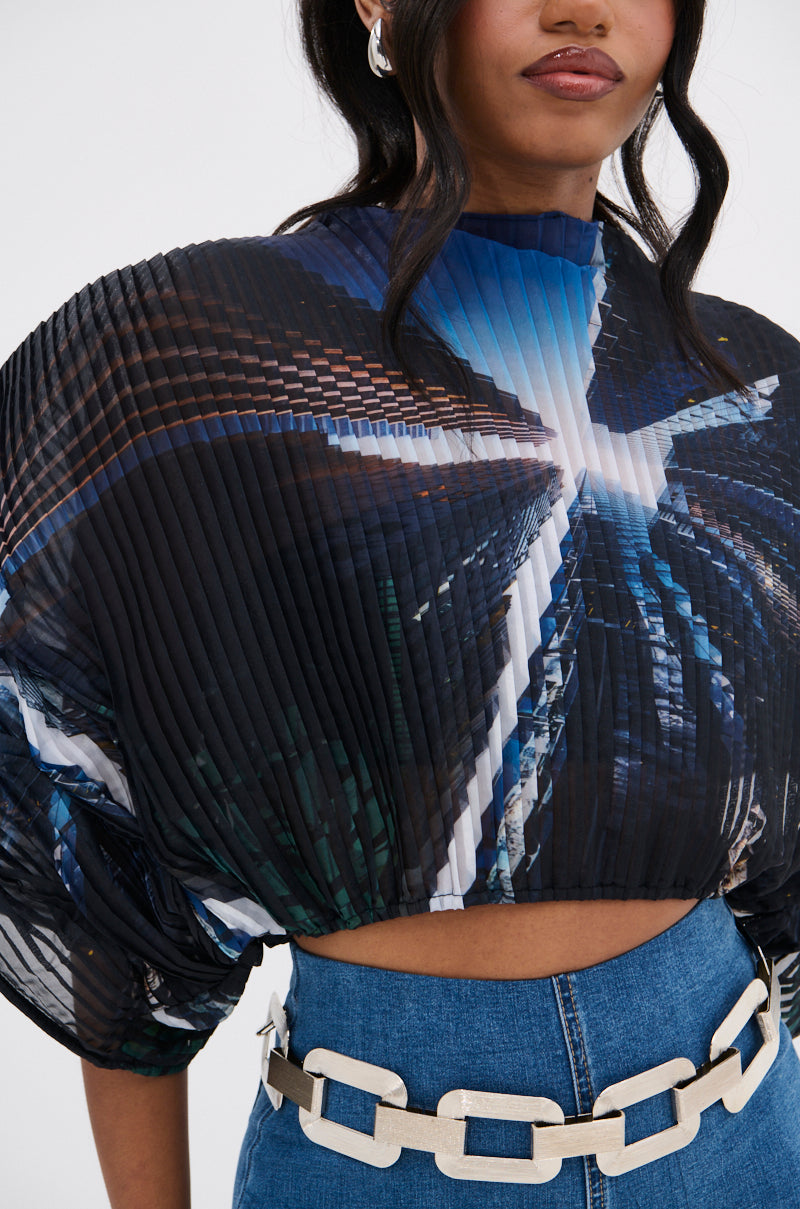 A woman wears the GALAXY BLOUSE—pleated, avant-garde with batwing sleeves and a bold abstract blue and white pattern—paired with high-waisted blue jeans and a chunky white chain belt. Only her lower face and torso are visible.