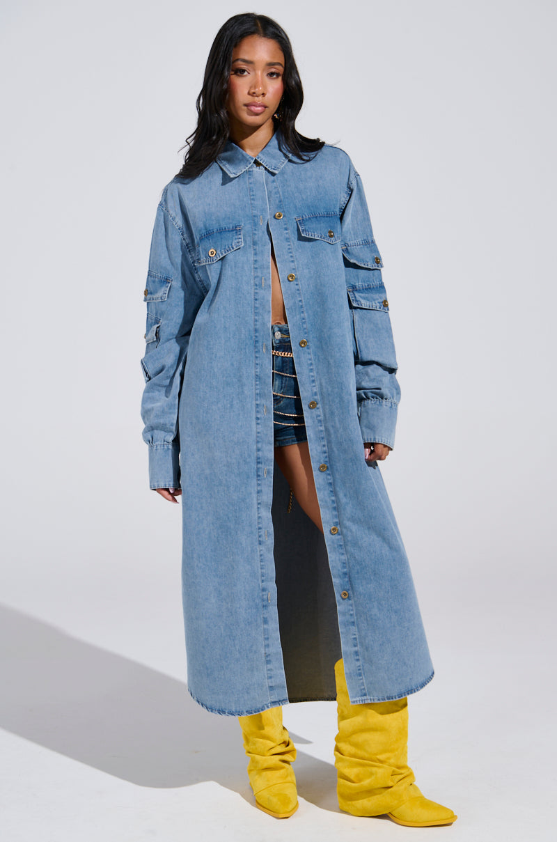 A woman confidently models the Comfort Queen Chambray Long Duster, featuring button details and layered sleeves, paired with shorts and striking yellow knee-high boots against a simple backdrop, embodying denim's timeless appeal.