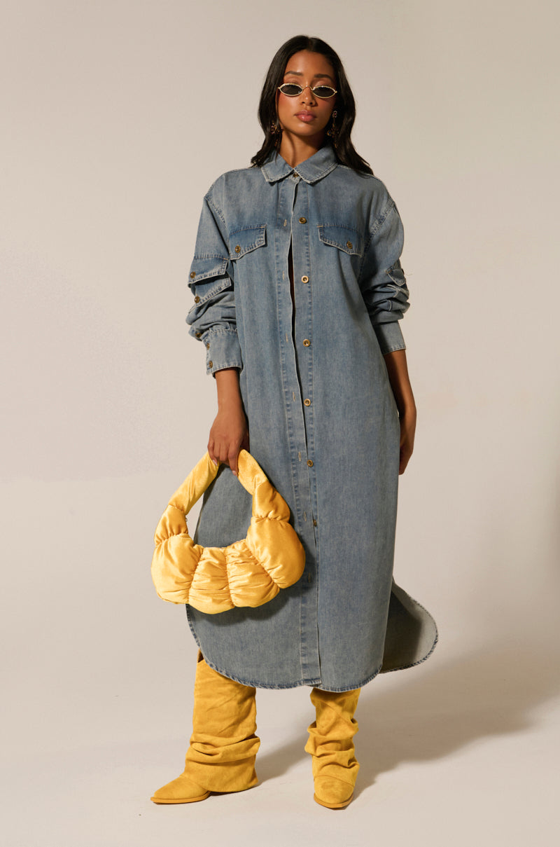 Wearing a COMFORT QUEEN CHAMBRAY LONG DUSTER, yellow knee-high boots, and a matching padded bag, they exude confidence. With sunglasses and dark wavy hair against a plain backdrop, they effortlessly showcase the chic appeal of denim on denim.
