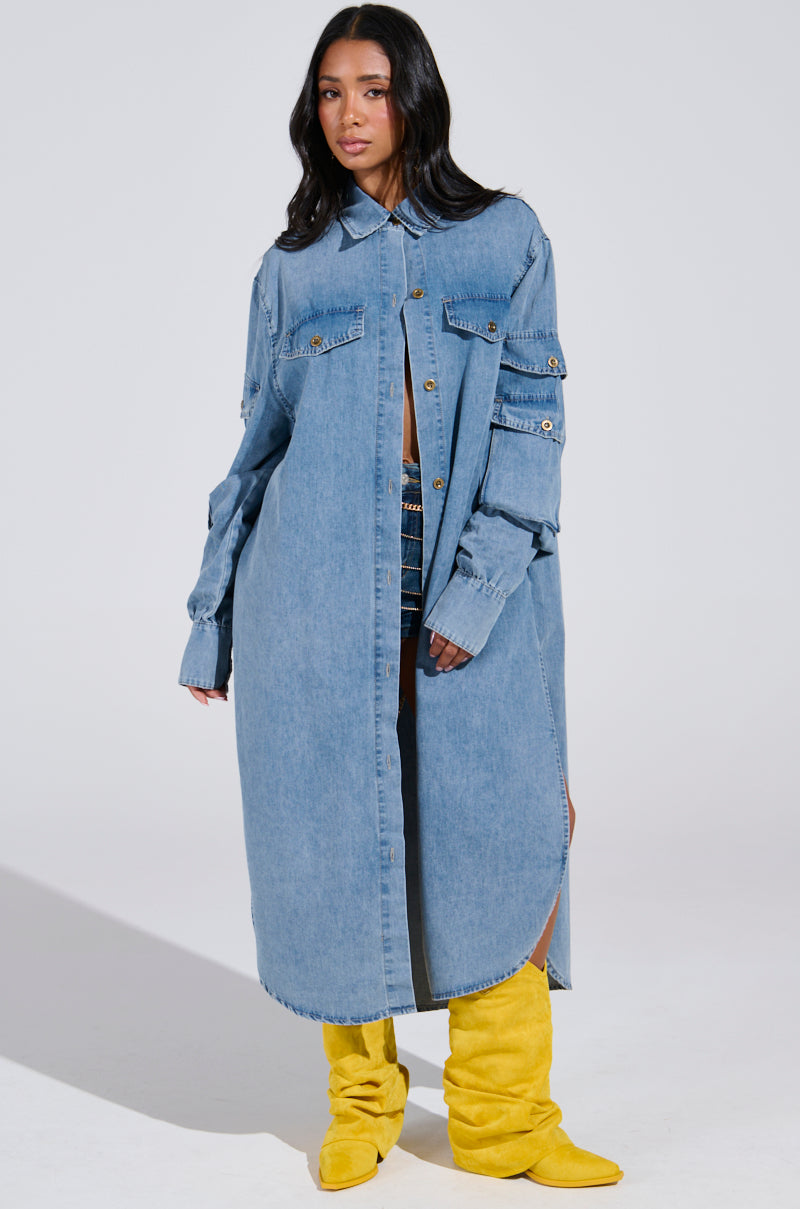 A person confidently stands in a COMFORT QUEEN CHAMBRAY LONG DUSTER with buttons, layered over a denim outfit with bright yellow slouchy boots. The plain light grey background accentuates their chic denim on denim style.
