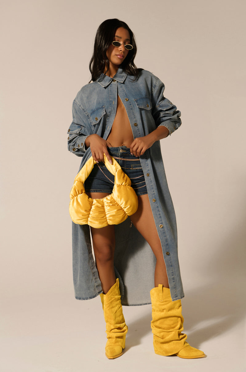 Wearing the COMFORT QUEEN CHAMBRAY LONG DUSTER and shorts, a woman radiates confidence. She pairs them with vibrant yellow boots, a matching bag, and round sunglasses that accent her long, dark hair for a blend of casual flair and denim chic, achieving an effortlessly stylish look.