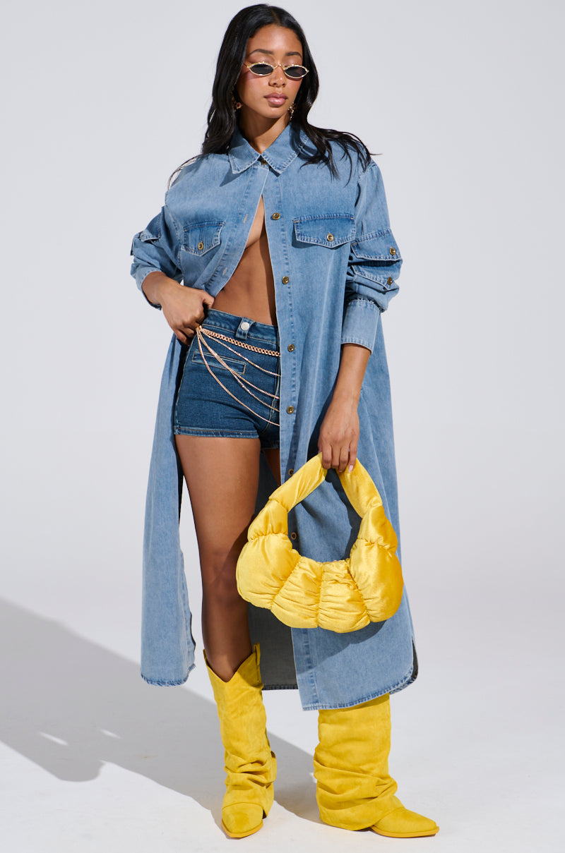 A woman confidently poses in the COMFORT QUEEN CHAMBRAY LONG DUSTER over denim shorts, embracing the denim-on-denim trend. She accessorizes with chain belts, yellow boots, a matching handbag, sunglasses, and flowing long hair against a plain background.
