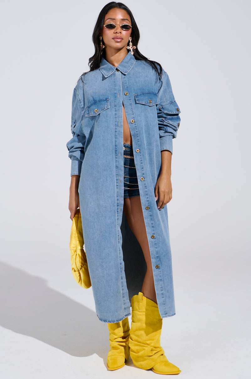 Wearing the COMFORT QUEEN CHAMBRAY LONG DUSTER over dark shorts and a top, paired with bright yellow boots and bag, a person poses confidently. With long hair, sunglasses, and hoop earrings against a simple backdrop, they nail the denim-on-denim look.
