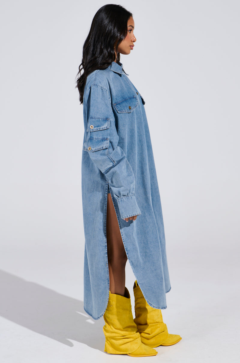 A person stands in profile wearing the COMFORT QUEEN CHAMBRAY LONG DUSTER with rolled sleeves and side slits, paired with vibrant yellow slouchy boots. The light-wash denim contrasts strikingly against the plain, neutral background.