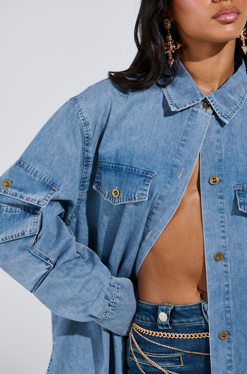 A person rocks a denim-on-denim look with a partially unbuttoned COMFORT QUEEN CHAMBRAY LONG DUSTER, revealing a bare midriff. They're adorned with ornate dangling earrings and a gold chain belt, with the photo cropped at the shoulders and waist.