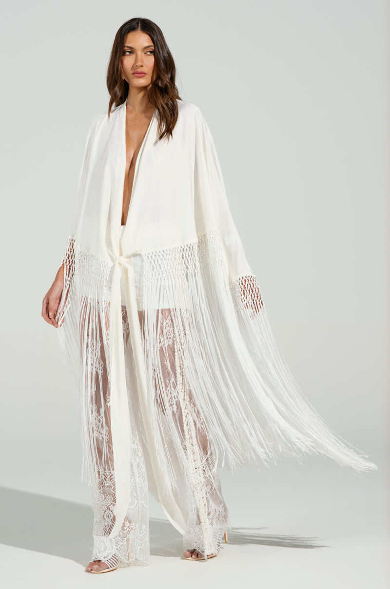 A woman models a flowing white ensemble featuring the "ON TO SOMETHING GOOD FRINGE TIE FRONT BLOUSE," paired with sheer lace pants. The belted top enhances the elegant and ethereal look, while tonal fringe trim adds whimsy against a neutral backdrop.