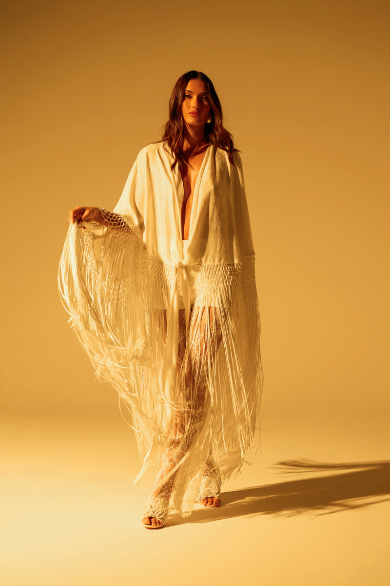 A woman stands confidently, her long hair cascading over the ON TO SOMETHING GOOD FRINGE TIE FRONT BLOUSE with tonal fringe trim and floral patterns. Set against a warm yellowish backdrop, the fabric elegantly falls to the floor, creating a dynamic and textured look.