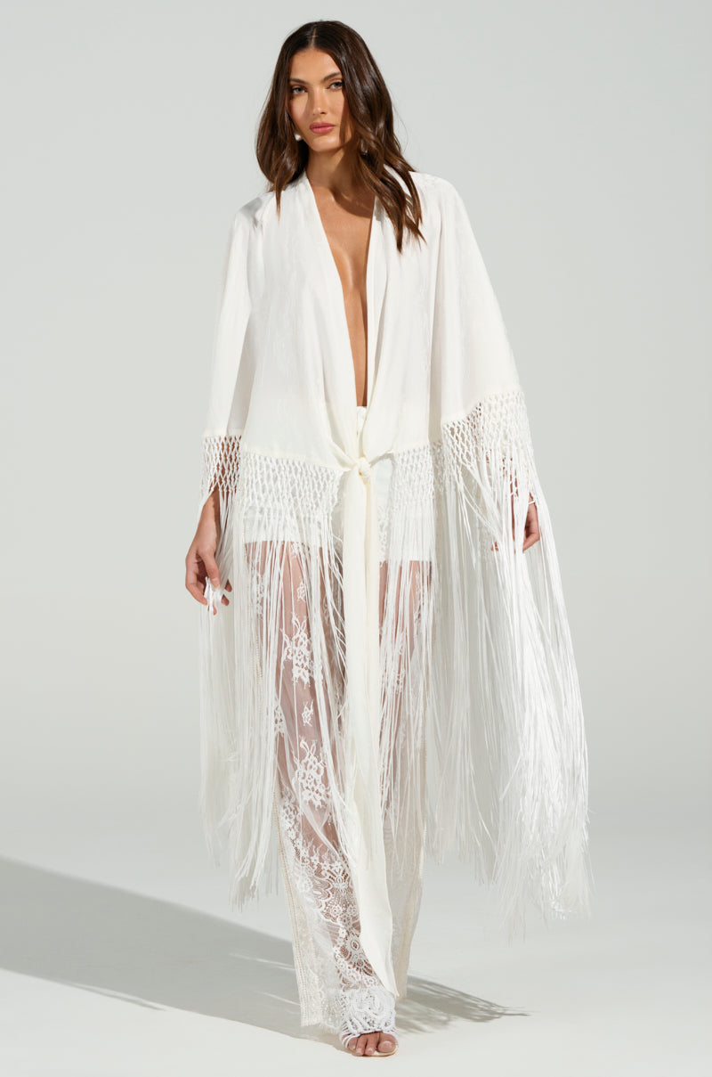 A woman poses in the ON TO SOMETHING GOOD FRINGE TIE FRONT BLOUSE, a sheer white kimono with long tonal fringe trim and intricate lace patterns. She pairs it elegantly with lace pants against a plain light gray backdrop.