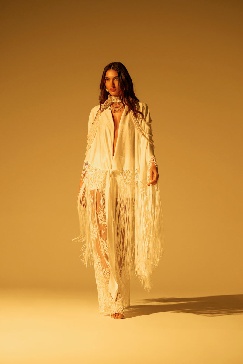 A woman stands confidently against a golden background, wearing the ON TO SOMETHING GOOD FRINGE TIE FRONT BLOUSE with a floral pattern and tonal fringe trim. Her loose hair and neutral makeup complement her relaxed yet strong pose, exuding effortless grace.