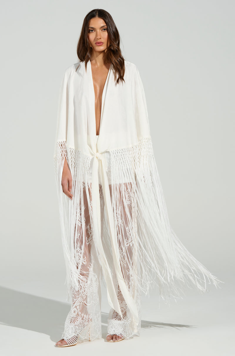 A woman in the ON TO SOMETHING GOOD FRINGE TIE FRONT BLOUSE, featuring white fringe and lace detailing on the sleeves and pants. This open-front, waist-tied blouse showcases a flowing, sheer design with a subtle floral pattern, set against a neutral background.