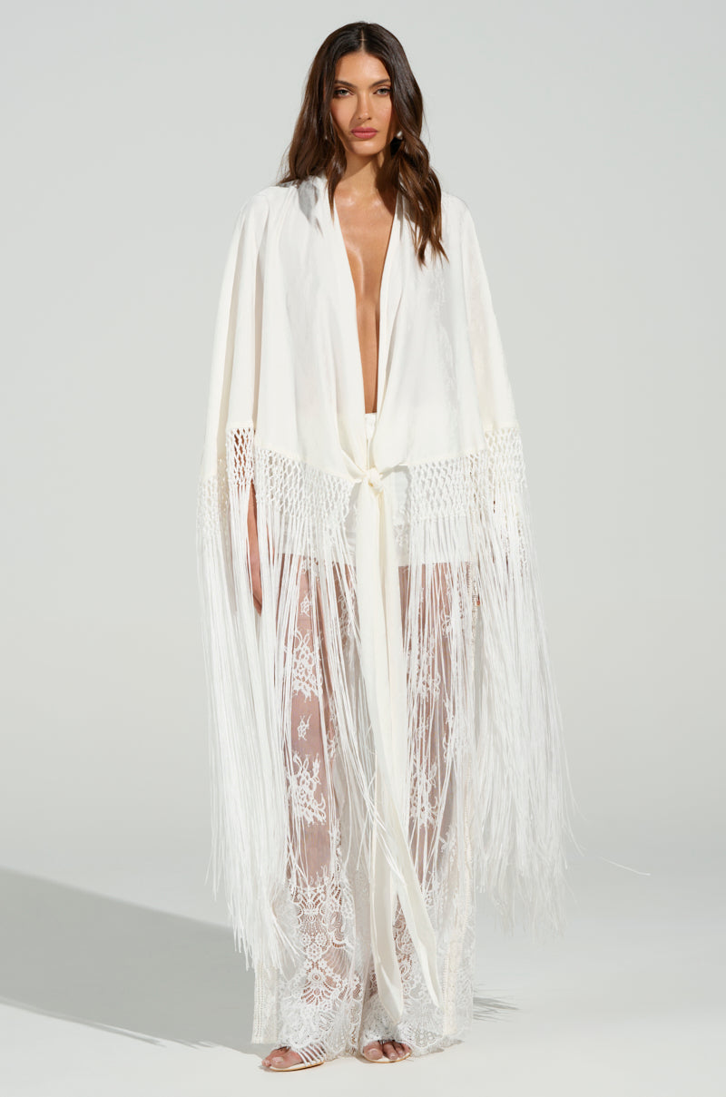 A woman stands wearing the "ON TO SOMETHING GOOD FRINGE TIE FRONT BLOUSE," a long, flowing sheer white garment with intricate lace and tonal fringe trim. She has long, wavy hair against a plain background.