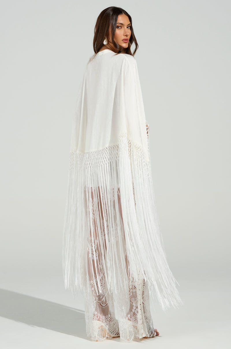 A woman elegantly poses in a flowing, lace-toned "On to Something Good Fringe Tie Front Blouse," her brunette hair mirroring the blouse's draped fringe. The neutral backdrop enhances her silhouette.