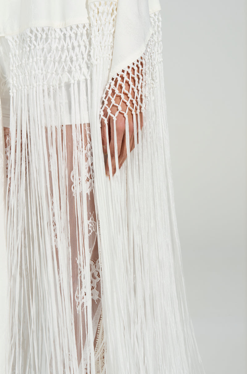 A person wears a white lace garment featuring tonal fringe trim and intricate macramé. The fabric drapes elegantly, echoing the allure of the "ON TO SOMETHING GOOD FRINGE TIE FRONT BLOUSE" against a neutral backdrop.
