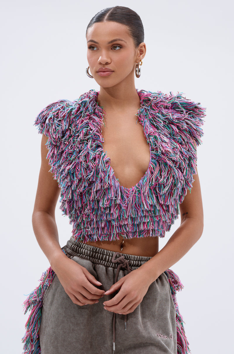 A woman stands against a plain background wearing THE ONE FRINGE BLOUSE—a sleeveless, deep V-neck multicolor yarn top featuring pink, blue, and purple fringe—paired with loose gray loungewear pants, hoop earrings, and her hair tied back.
